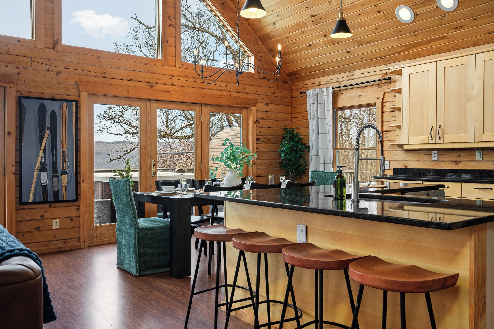 A wooden cabin interior with a dining area, bar stools, a kitchen with a black countertop, and a large window overlooking a landscape.