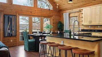 A wooden cabin interior with a dining area, bar stools, a kitchen with a black countertop, and a large window overlooking a landscape.