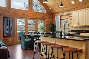 Interior de una cabaña de madera con comedor, taburetes de bar, cocina con encimera negra y un gran ventanal con vistas al paisaje.