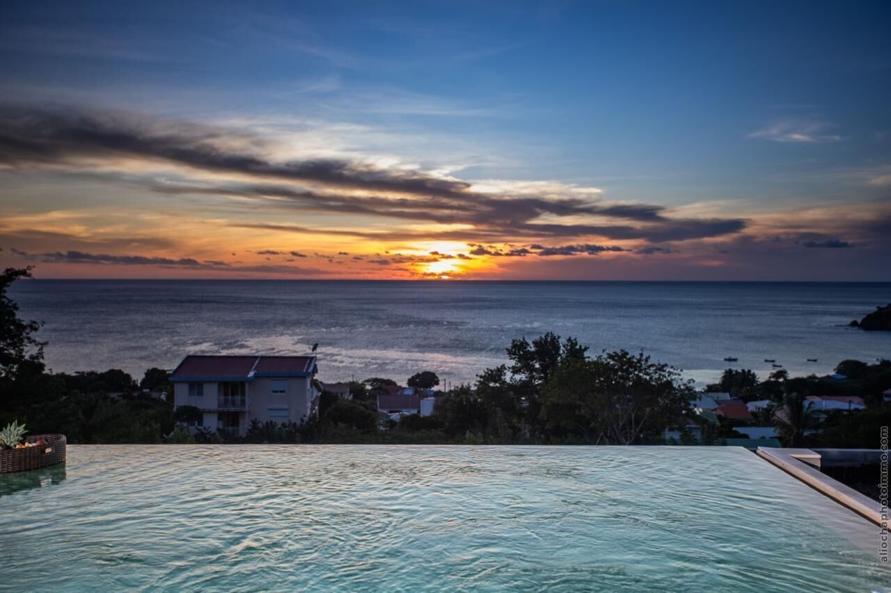 Vista al tramonto da una piscina a sfioro con vista sull'oceano, con case e alberi sullo sfondo.