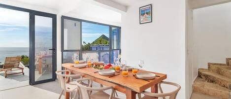 Een eetgedeelte met een houten tafel voor zes personen, uitzicht op zee, en een trap naar een hoger gelegen niveau.