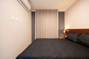 A hotel room with a bed, a wall-mounted air conditioner, a light switch, and a window with vertical blinds. - Estúdio Premium Vila Olímpia -ap1901 (Sao Paulo)