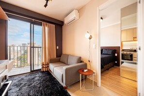 A modern hotel room with a sofa, a small round table, a lamp, a bed, and a view of the cityscape through large windows. - Estúdio Premium Vila Olímpia -ap1901 (Sao Paulo)