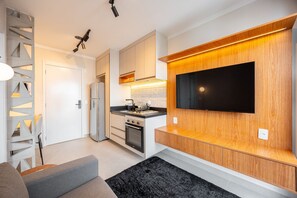 A modern kitchen with a built-in oven, a flat-screen TV mounted on the wall, and a grey sofa in the living area. - Estúdio Premium Vila Olímpia -ap1901 (Sao Paulo)