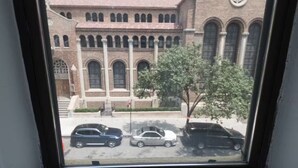 Blick aus dem Fenster auf ein Gebäude mit rotem Dach, Rundbogenfenstern und einem kreisförmigen Fenster an der Fassade.
