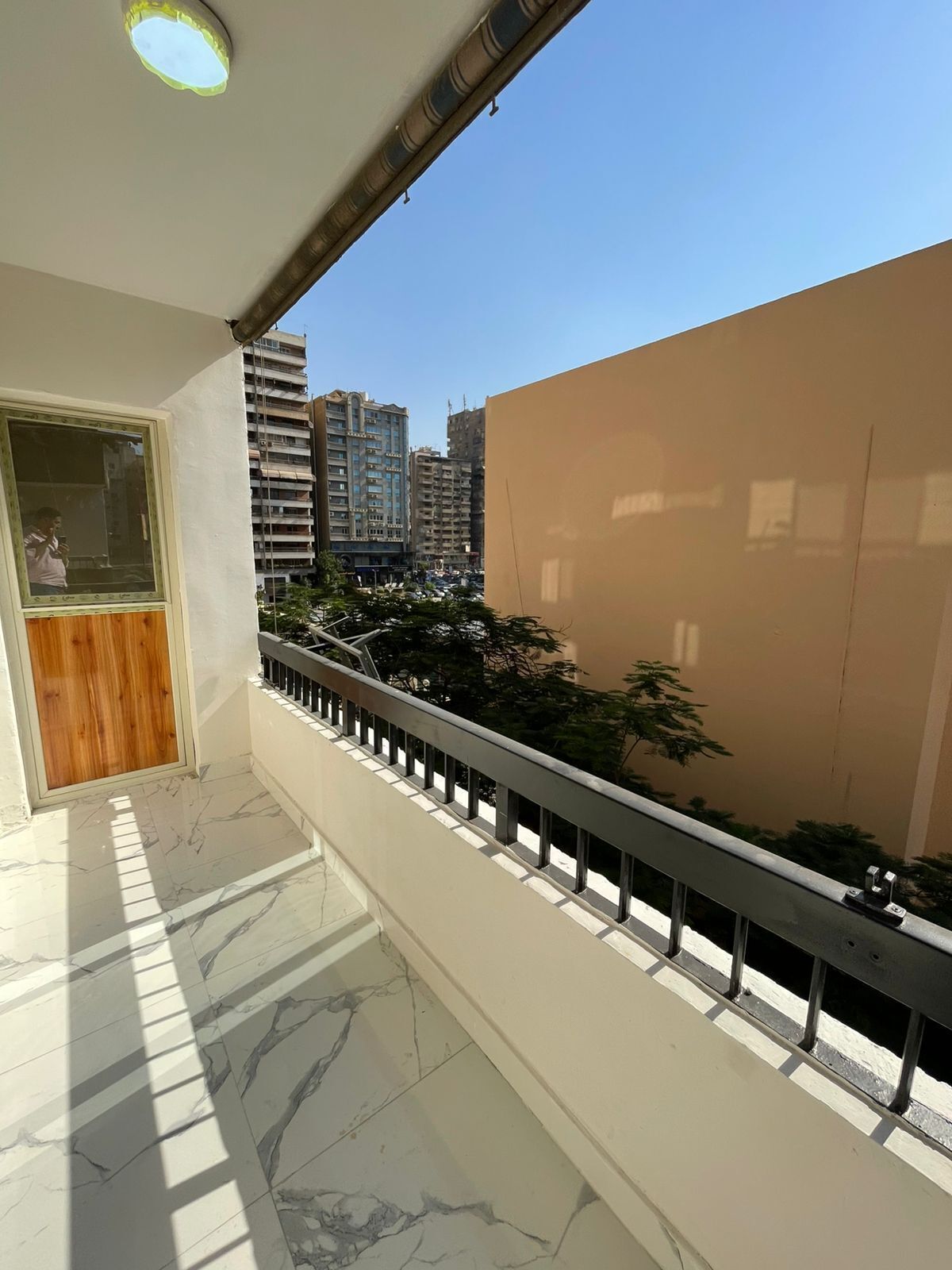 A balcony with a marble floor, a wooden door, and a view of a cityscape with high-rise buildings.