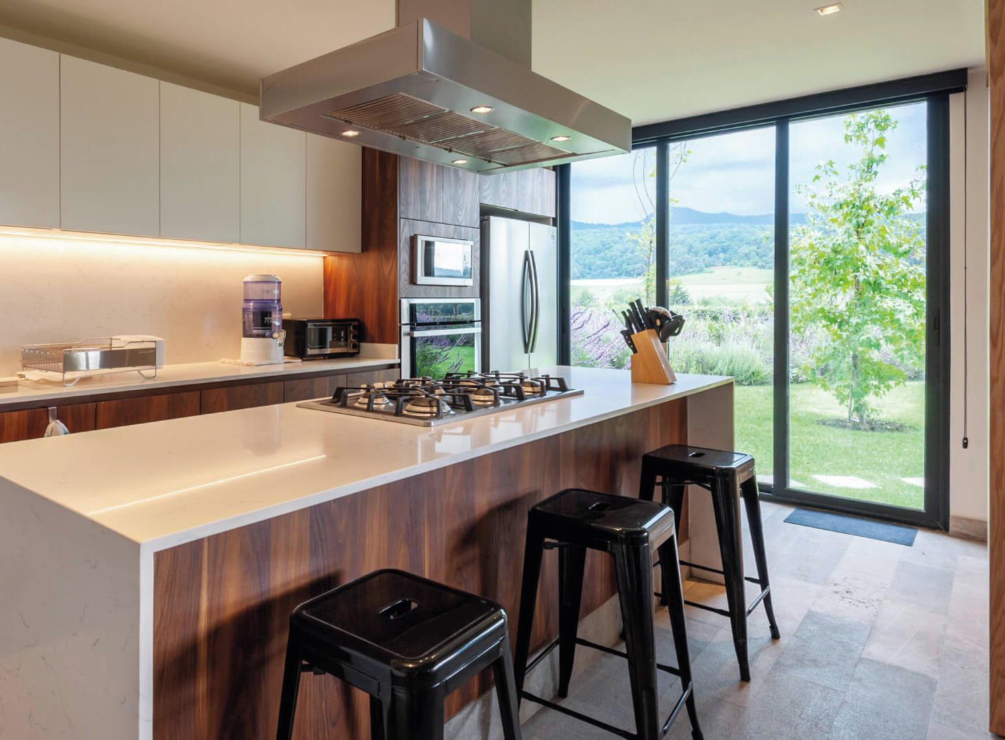 A modern kitchen with a large island, stainless steel appliances, and a view of a green landscape through large windows.