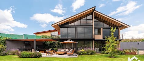 A modern two-story house with a large glass facade, a covered patio with outdoor furniture, and a well-maintained garden.