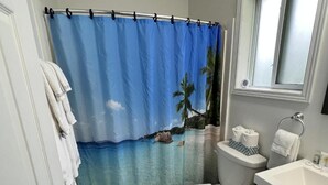 A bathroom with a blue shower curtain depicting a beach scene, a toilet, a sink, and a towel rack. - Jacuzzi Cabin - 25 min to Blue Mountain,  3 min Walk to Beach (Wasaga Beach)