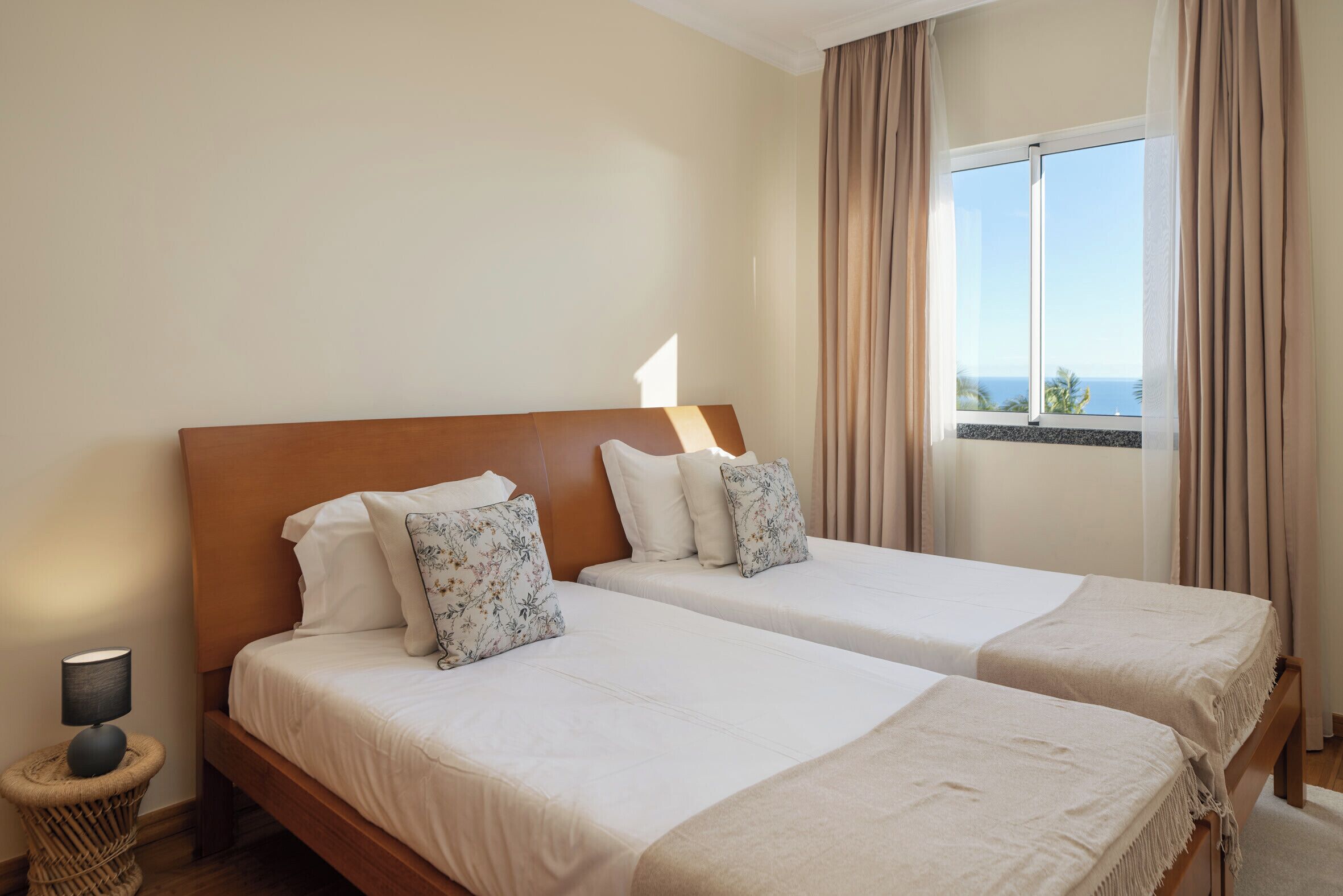 A hotel room with two beds, a bedside lamp, a wicker side table, and a window with a view of the sea.