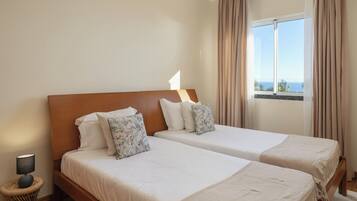 A hotel room with two beds, a bedside lamp, a wicker side table, and a window with a view of the sea.