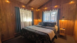 A cozy bedroom with a wooden ceiling, a large bed with patterned bedding, two windows with sheer curtains, and a bedside table with a clock. - Owl's Nest at Feather Nest Inn (Grand Marais)