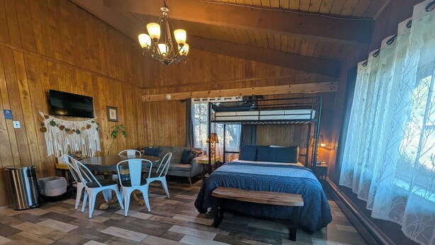 A cozy room with a wooden floor, a bed with a blue bedspread, a dining table with chairs, and a chandelier. - Owl's Nest at Feather Nest Inn (Grand Marais)