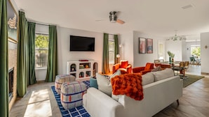 A modern living room with a sofa, ottoman, and a flat-screen TV. There is a dining area with a table and chairs. - Playtime Bonanza House - Game Room & Playground (Austin)