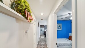 Un couloir étroit avec un plancher en bois, un mur blanc orné d’une plante, et une chambre bleue avec un lit et un tableau accroché au mur.