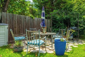Une terrasse à l’arrière de la maison, avec une clôture en bois, une table entourée de chaises, un parasol bleu et des plantes en pot.