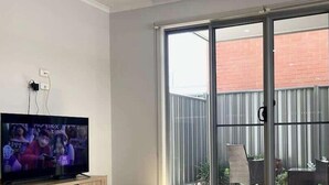 Un salon équipé d’un téléviseur, d’une porte-fenêtre coulissante donnant sur un balcon et d’un fauteuil en osier.