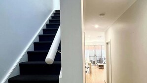 Un intérieur moderne avec un plancher en bois, un escalier aux marches sombres et un couloir lumineux menant à une pièce équipée d’un bureau et d’une chaise.
