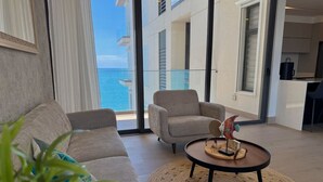 A modern living room with a sofa, armchairs, and a coffee table with a decorative bird sculpture. A large window offers a view of the ocean and a building with balconies. - The O apartments (Mazatlán)