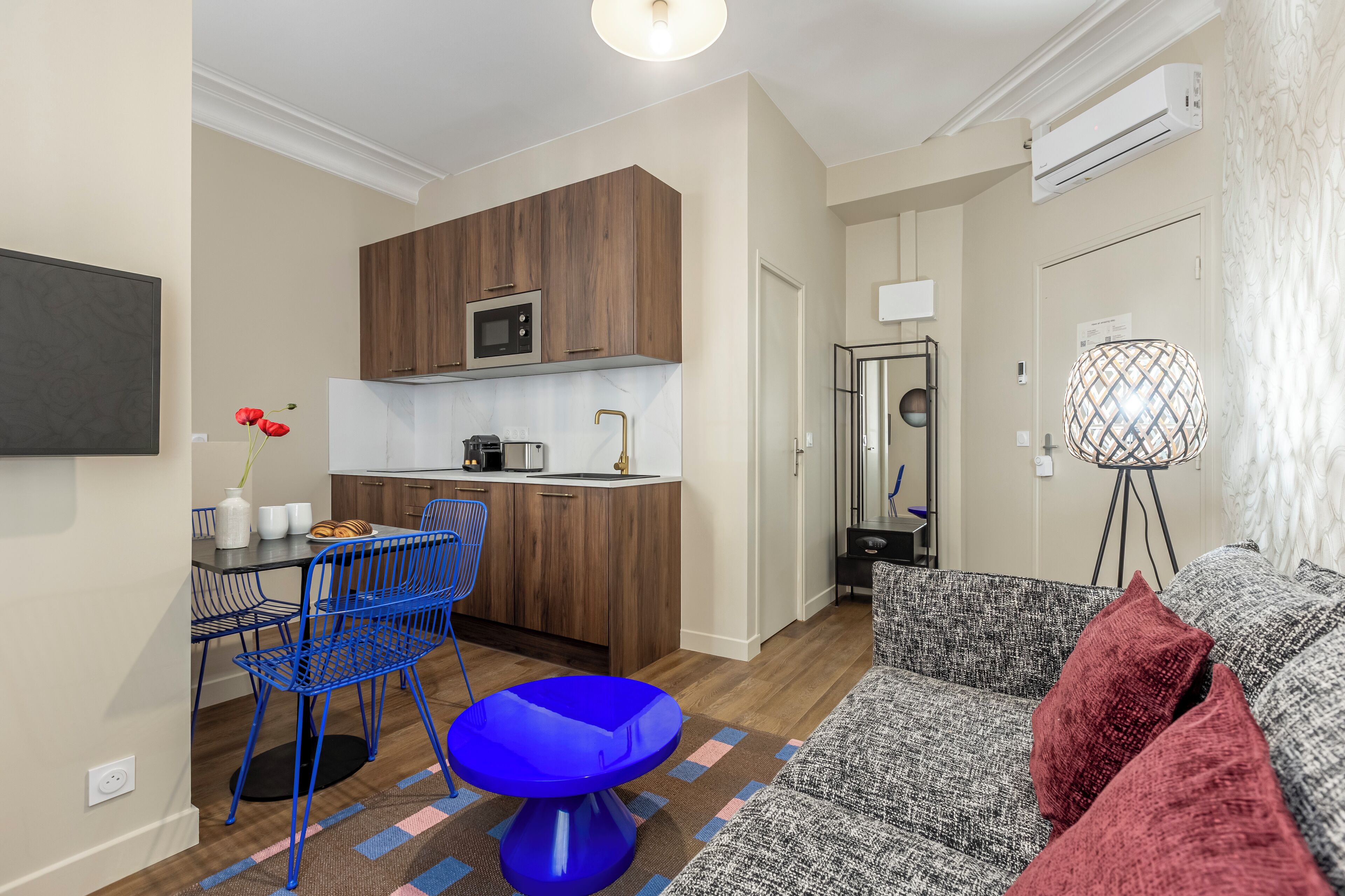 Un espace de vie compact comprenant un coin cuisine, un coin repas avec des chaises bleues, un fauteuil d’accent bleu, un tapis à motifs et un canapé gris avec des coussins rouges.