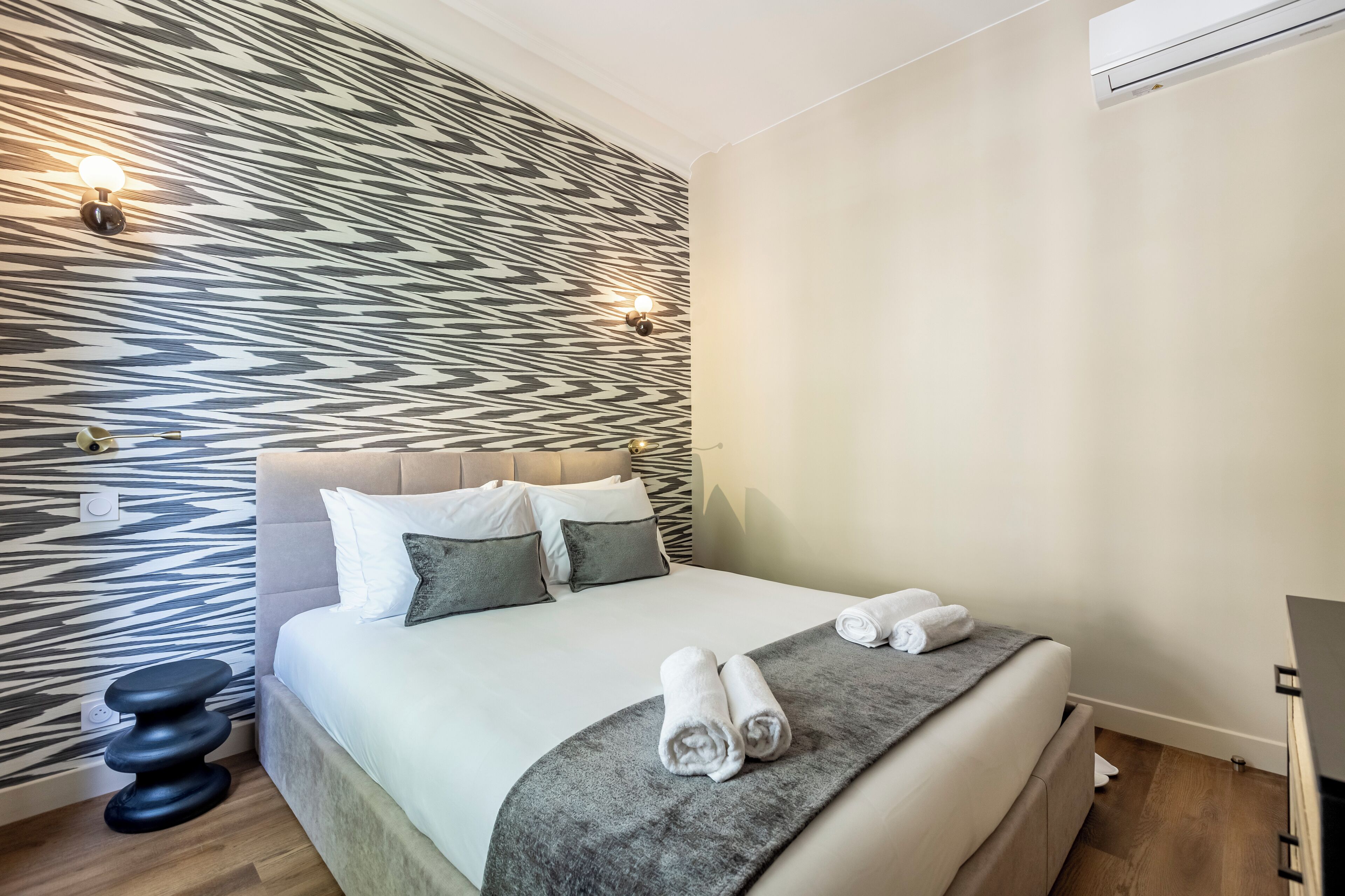 Ein modernes Hotelzimmer mit Wandpaneelen, einem Bett mit weißen Leinentüchern, zwei Kissen und einem Nachttisch mit Lampe.