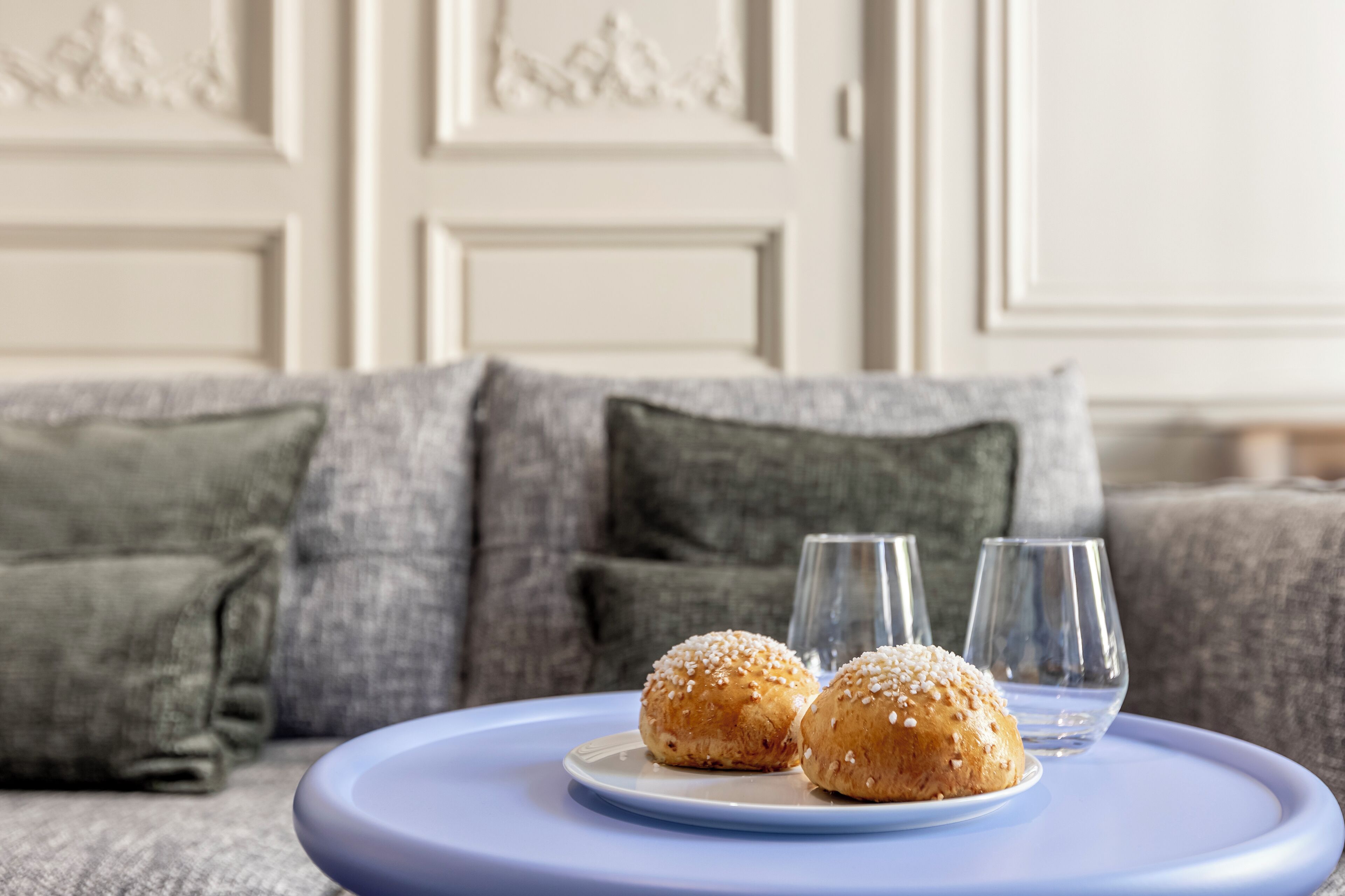Una mesa con dos pasteles y dos vasos sobre una bandeja azul, colocada sobre un sofá gris con almohadones decorativos.