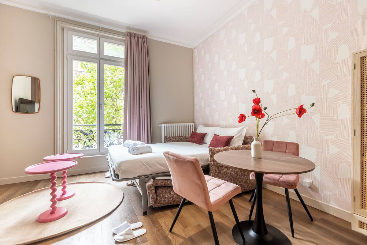 Una habitació d'hotel amb un llit, una taula de dinar amb cadires, una taula petita rosa i un mirall a la paret.