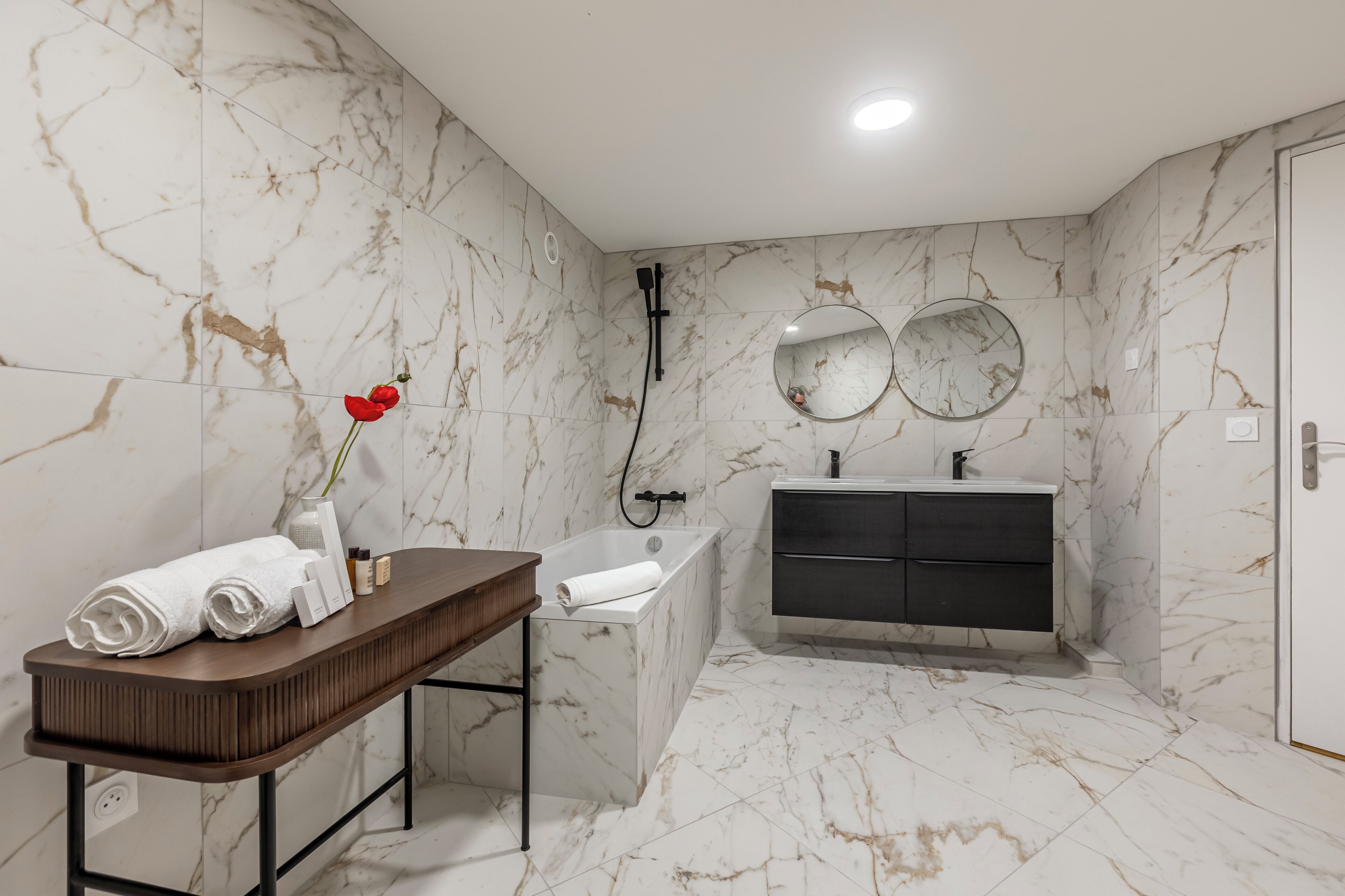 Un baño moderno con paredes de mármol, un mueble de tocador de madera, una bañera y un armario negro.