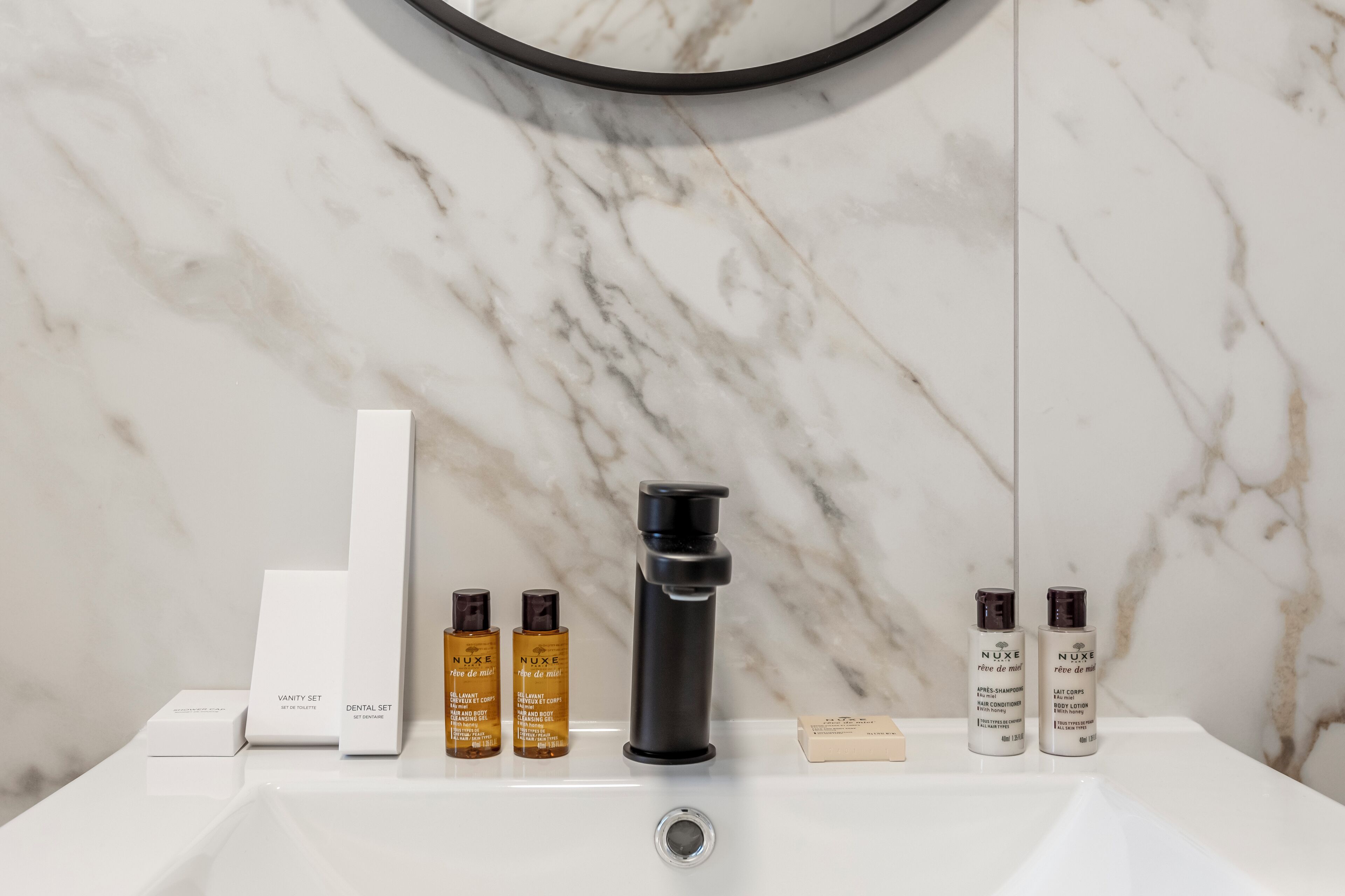 A bathroom with a marble sink, a mirror, and various toiletries including a vanity set, dental set, and body lotion.