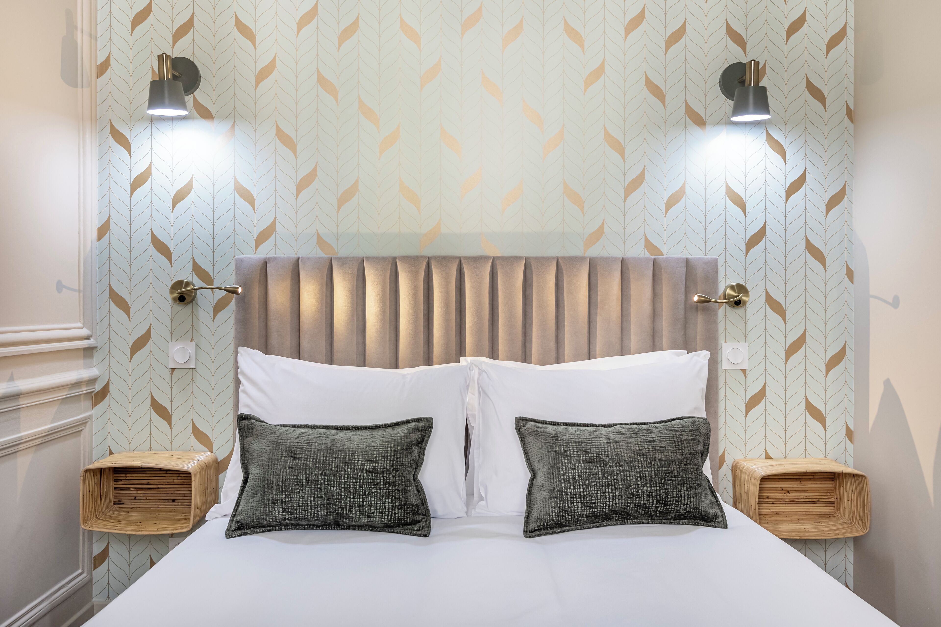 Quarto de hotel com uma cama, duas mesas de cabeceira, uma luminária na parede e papel de parede estampado.