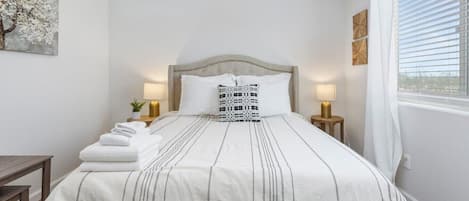 Una cama bien hecha con un edredón a rayas, dos lámparas de noche y un ventanal con persianas.