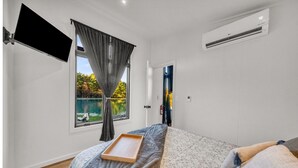 Un dormitorio con cama, televisor montado en la pared, ventana con cortinas, aire acondicionado y una puerta que da a otra habitación.