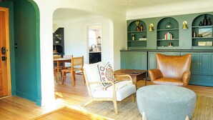A living room with a wooden floor, a green archway, a brown leather armchair, a white armchair with a patterned pillow, and a grey ottoman. - Cozy, Comfortable Home Base in Laurelhurst, Portland (Portland)