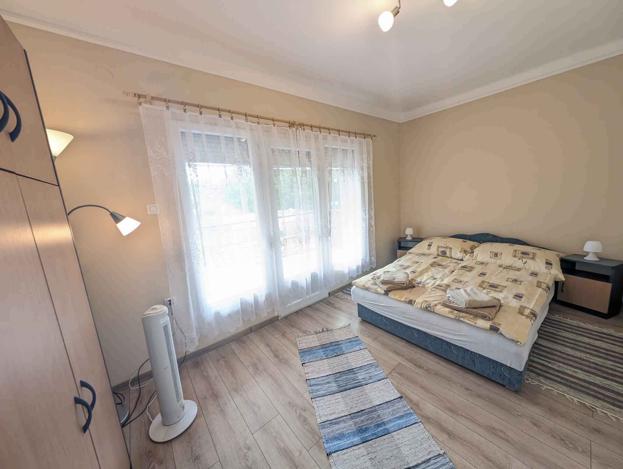 Une chambre à coucher avec un lit, des tables de chevet, un ventilateur sur pied et une fenêtre avec des rideaux.