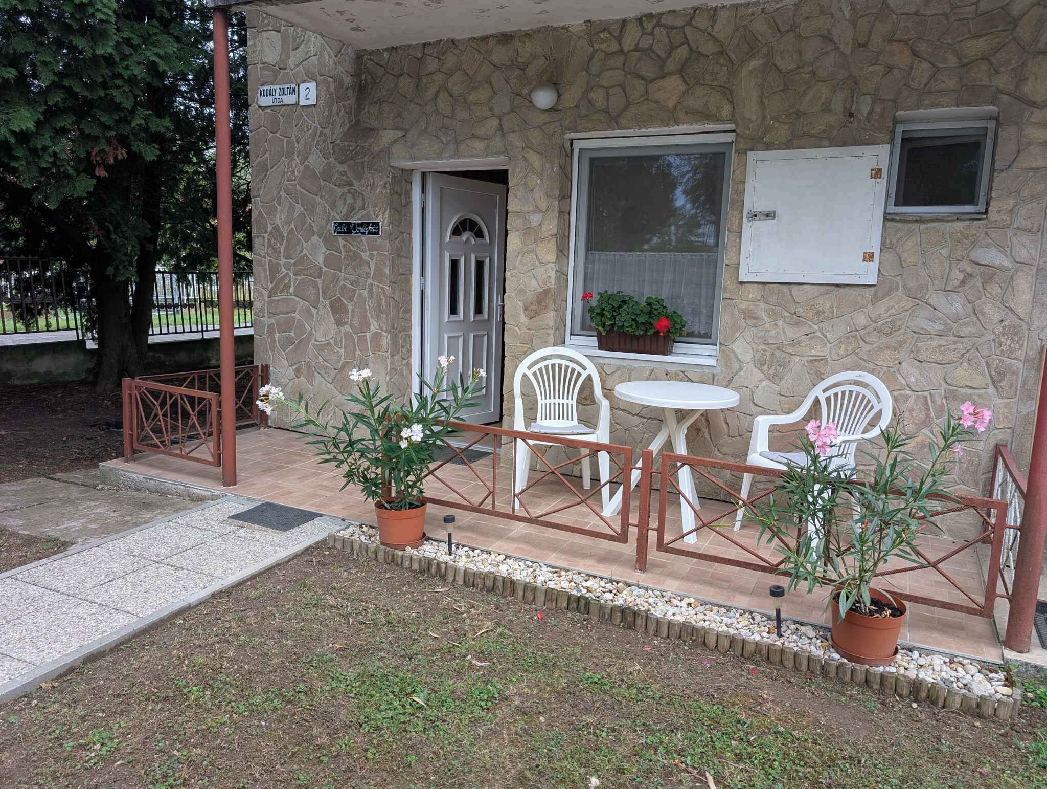 Un edificio in pietra con un piccolo patio, dotato di due sedie bianche e un tavolo, circondato da piante in vaso.