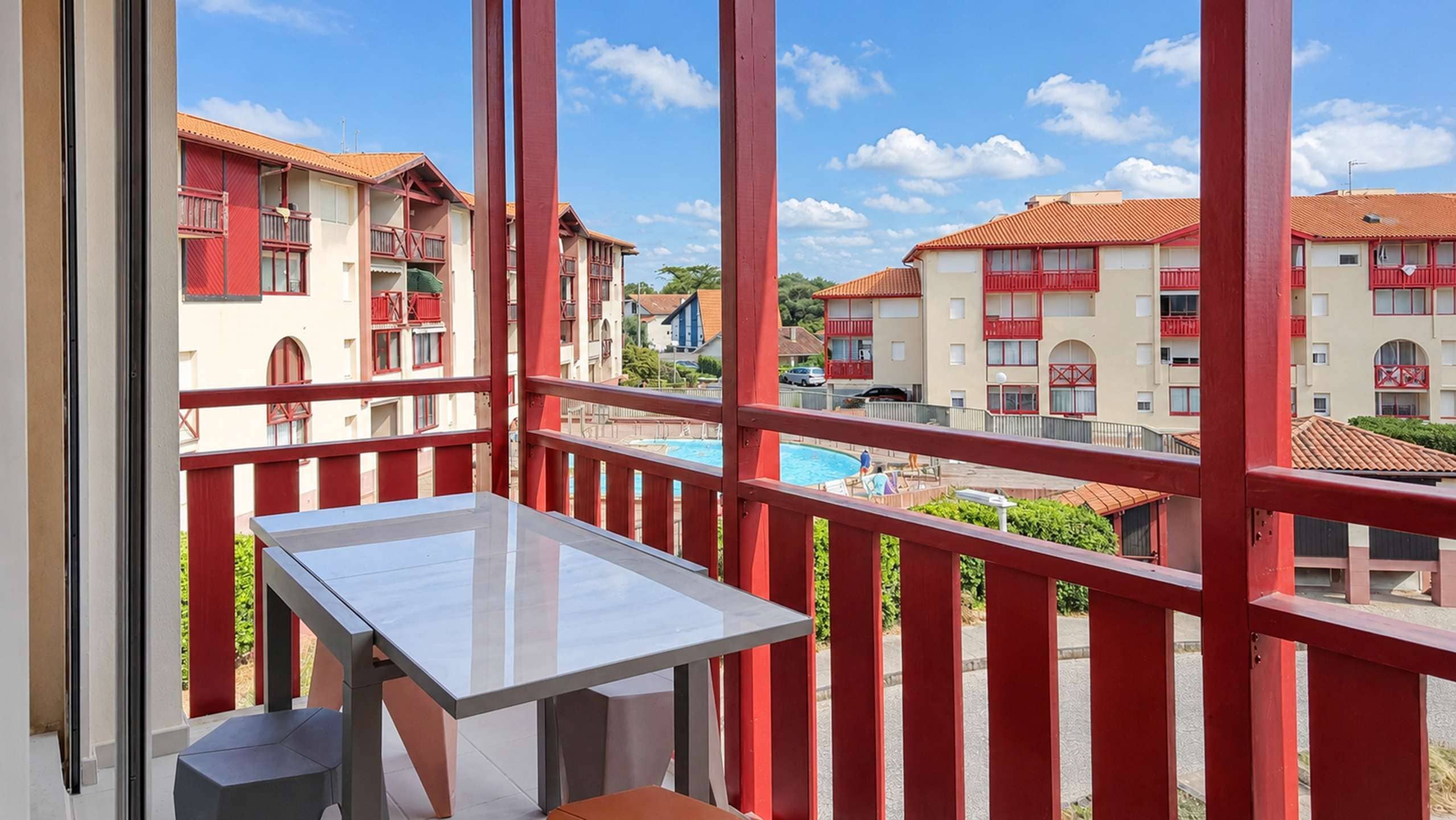 A balcony with a table and chairs, overlooking a swimming pool and buildings with red balconies.