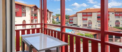 A balcony with a table and chairs, overlooking a swimming pool and buildings with red balconies.