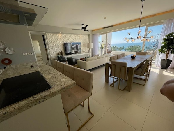 Uma sala de estar moderna com sofá, mesa de jantar e televisão. Há uma varanda com vista para o oceano.