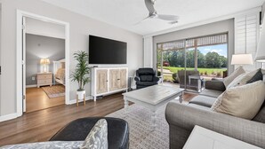 Un soggiorno moderno con una TV a schermo piatto, un ventilatore a soffitto e un'ampia finestra vista giardino.