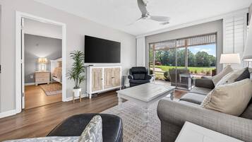 A modern living room with a flat-screen TV, a ceiling fan, and a large window offering a view of a garden.