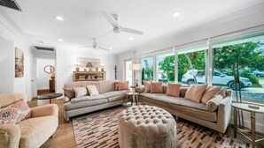 A spacious living room with a beige sofa, a round ottoman, and a coffee table. There are ceiling fans, a bookshelf, and a large window with a view of the street. - 633 Managed by Brampton Park (Boynton Beach)