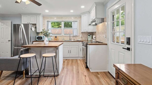 Una cocina moderna con armarios blancos, una isla de madera con taburetes, horno empotrado y ventilador de techo.