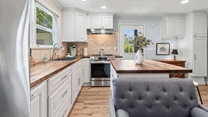 Una cocina con alacenas blancas, encimera de madera, horno empotrado y una silla tapizada en gris.