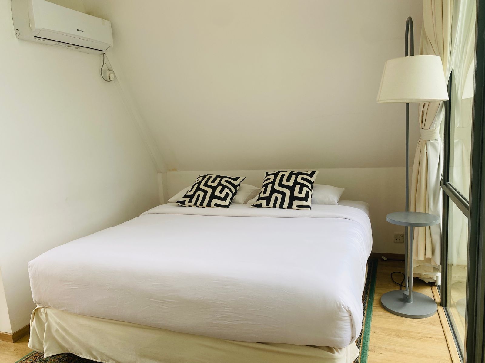 A neatly made bed with white bedding and two black and white patterned pillows, a standing lamp, and a window with curtains.