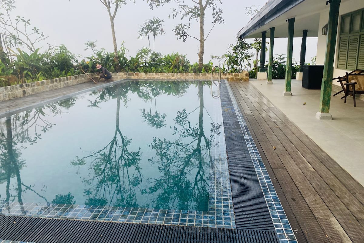 Une piscine dont le bord est revêtu de carreaux, entourée d’une terrasse en bois et d’un espace abrité avec des chaises.