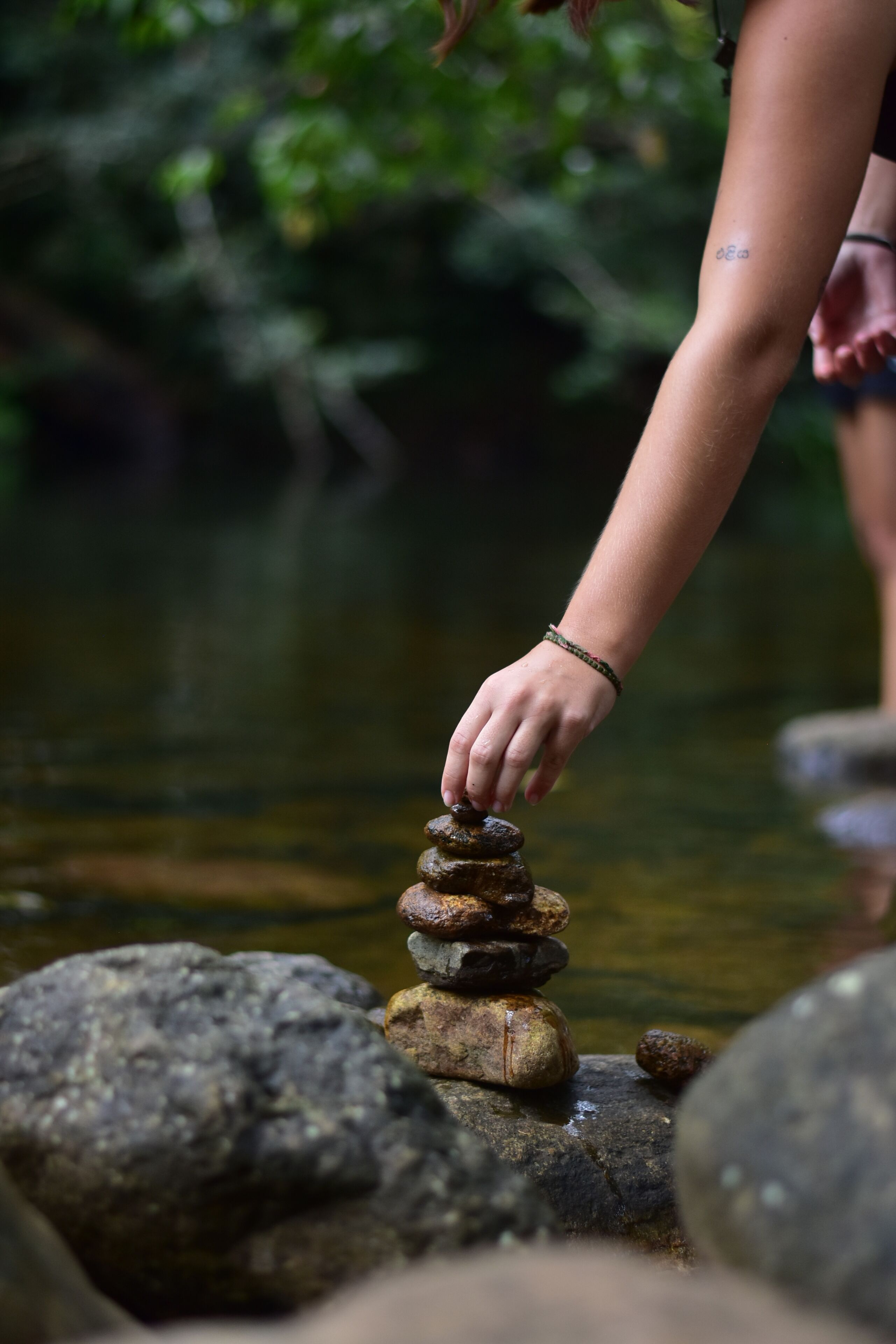 A hand balancing stones on a rock by a calm water body.