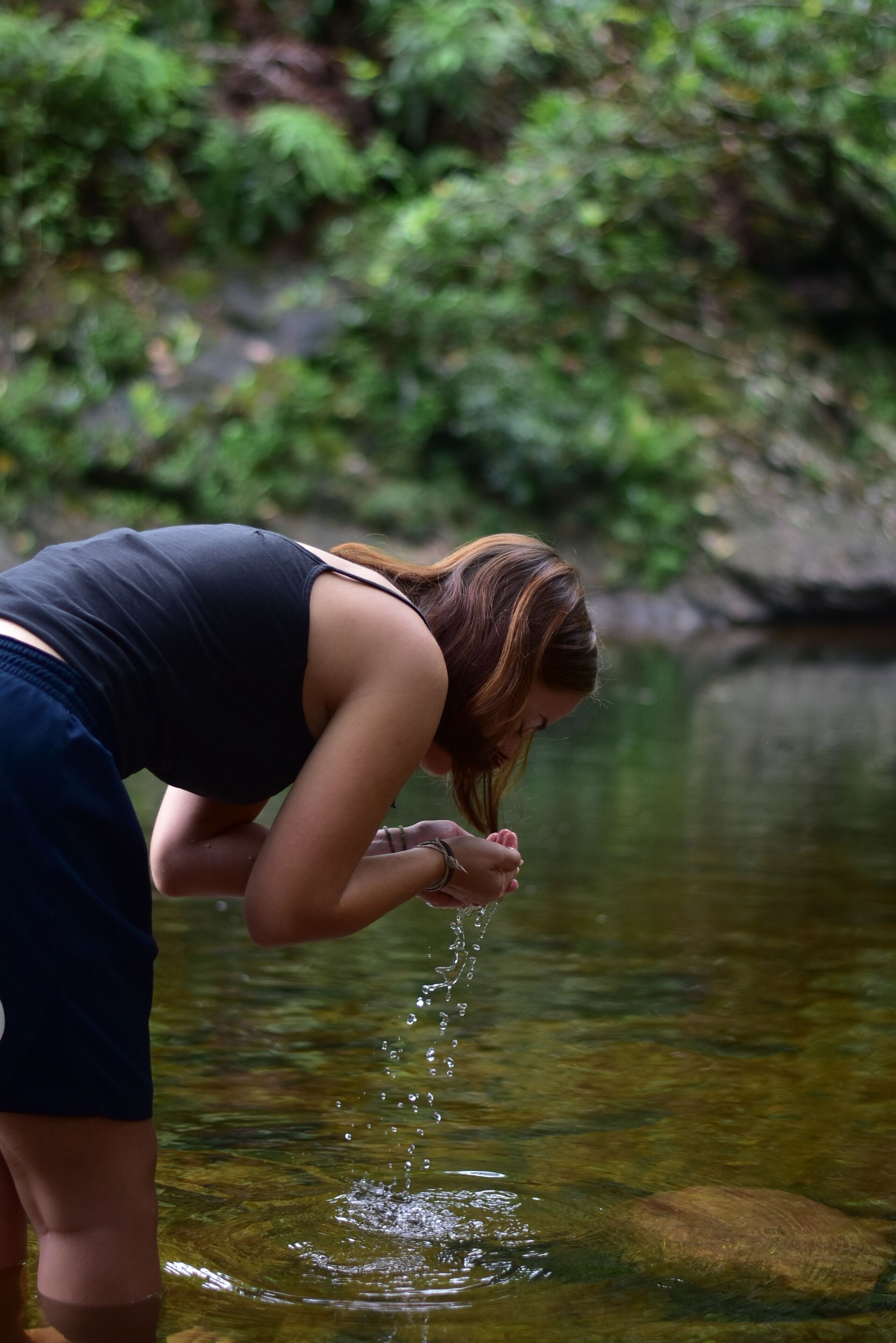 A person bending over a clear stream, with greenery in the background.