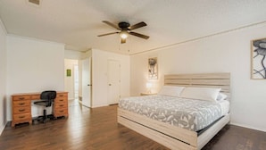 Quarto com cama, escrivaninha, cadeira, ventilador de teto e mesa de cabeceira com abajur.