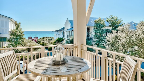 Una terrazza in legno con un tavolo rotondo e sedie, con vista sulla costa, edifici e alberi.