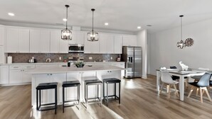 Cocina moderna con armarios blancos, una isla central con taburetes de bar, electrodomésticos de acero inoxidable e iluminación colgante.